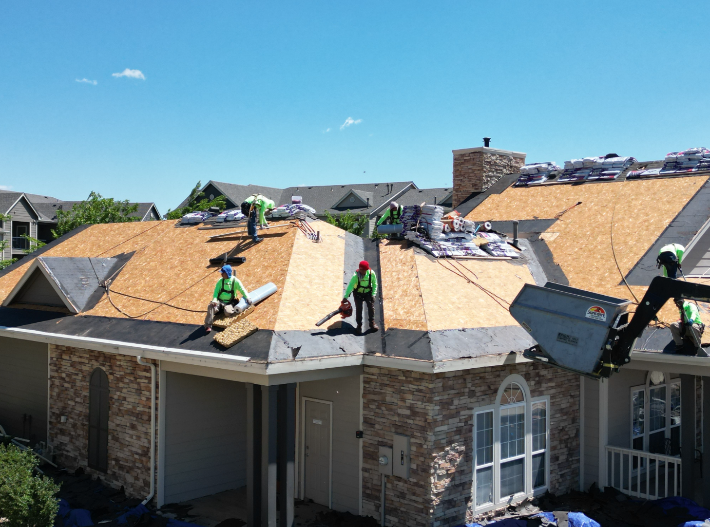 Aerial of Force 5 Roofing crew laying fresh decking and underlayment across a multifamily building, shingle bundles staged and dump trailer chute positioned