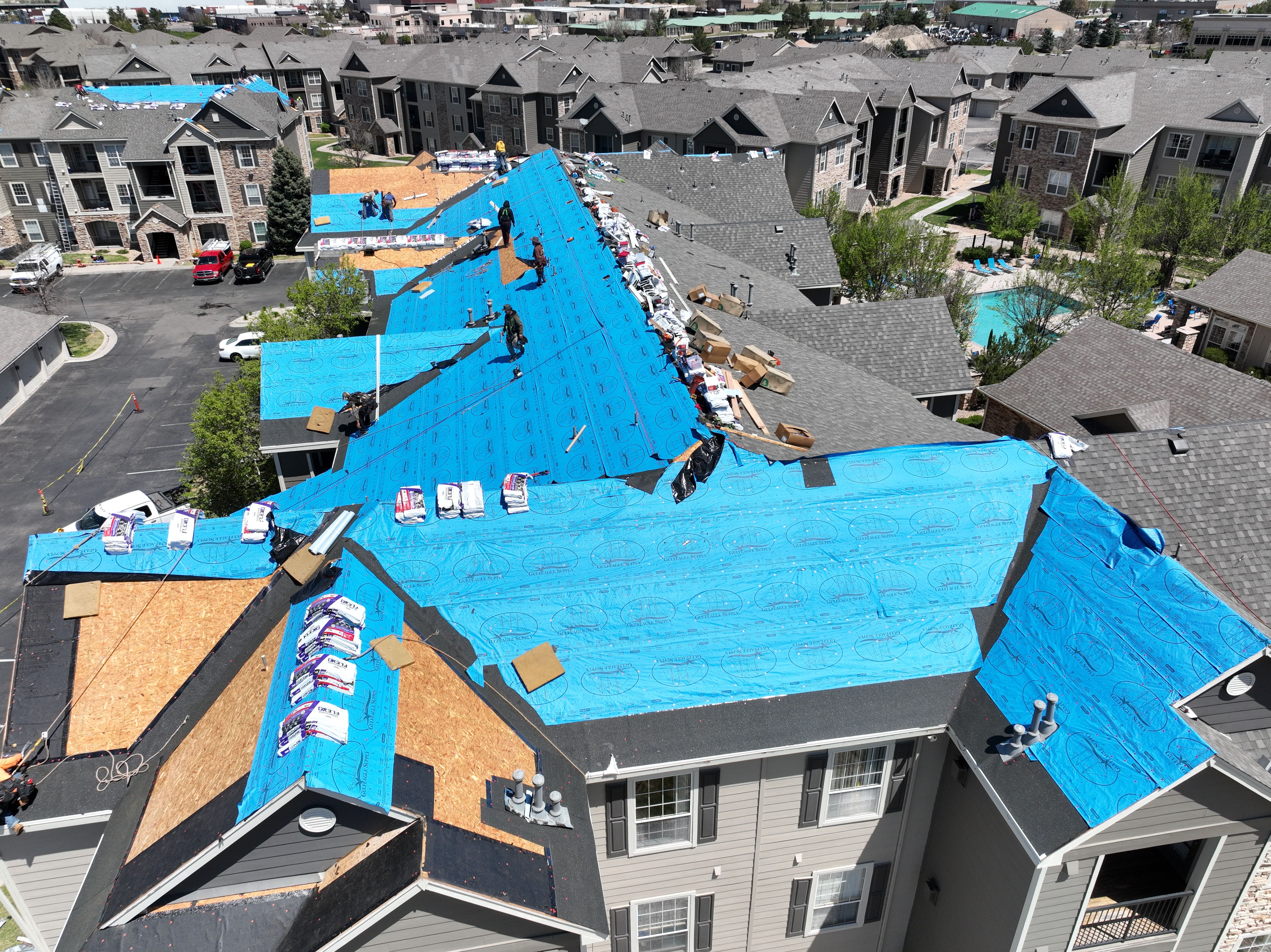 Aerial ridge-line view of Force 5 Roofing crew working a long multifamily building, with underlayment laid and shingle bundles staged the full length of the roof