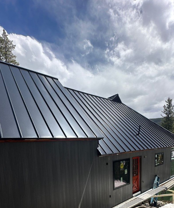 Close-up of a Force 5 Roofing standing seam metal roof installation on a dark-stained Summit County mountain home, vertical seams running up to a Colorado-blue sky, red entry door visible below