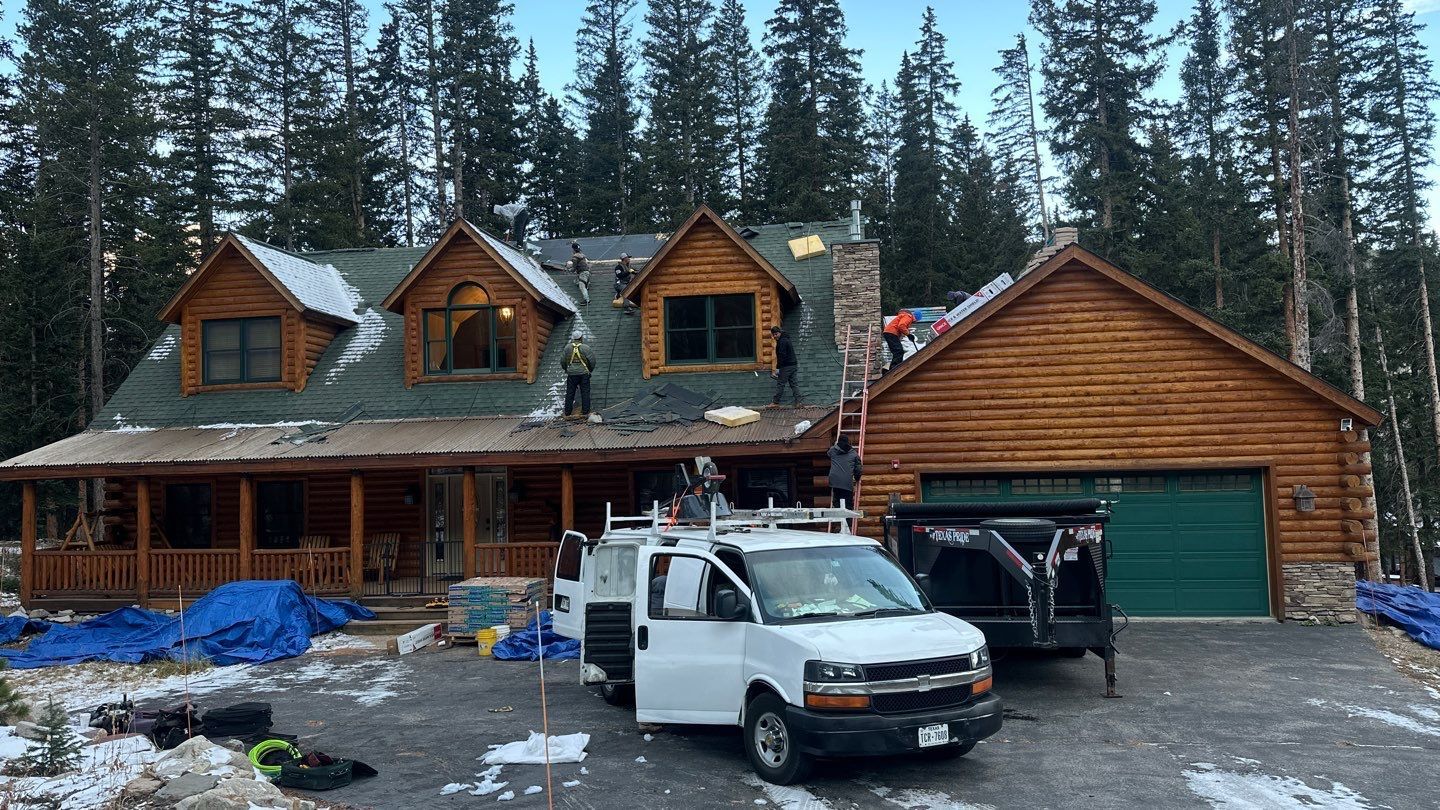 Force 5 Roofing crew installing a new roof on a log cabin in Summit County, CO — branded van and dump trailer staged in the driveway, surrounded by pines