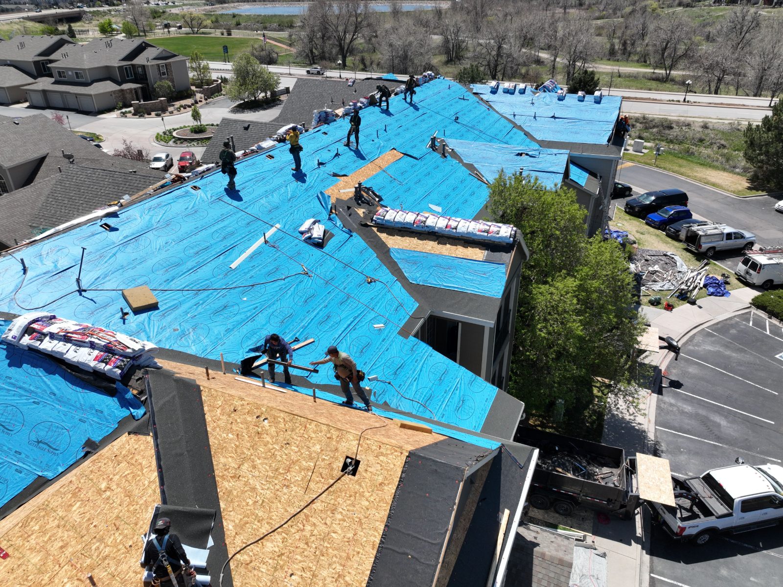 Aerial view of a Force 5 Roofing crew re-roofing a large multi-building complex — synthetic underlayment down, shingle bundles staged, dump trailer below
